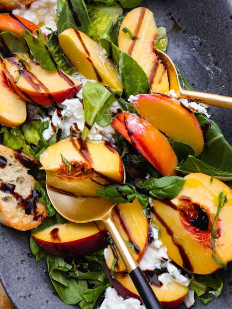 Close up view of peach burrata salad.  Golden and black serving spoons are placed in the salad.  Balsamic glaze drizzled on top and topped with crostini and fresh basil.