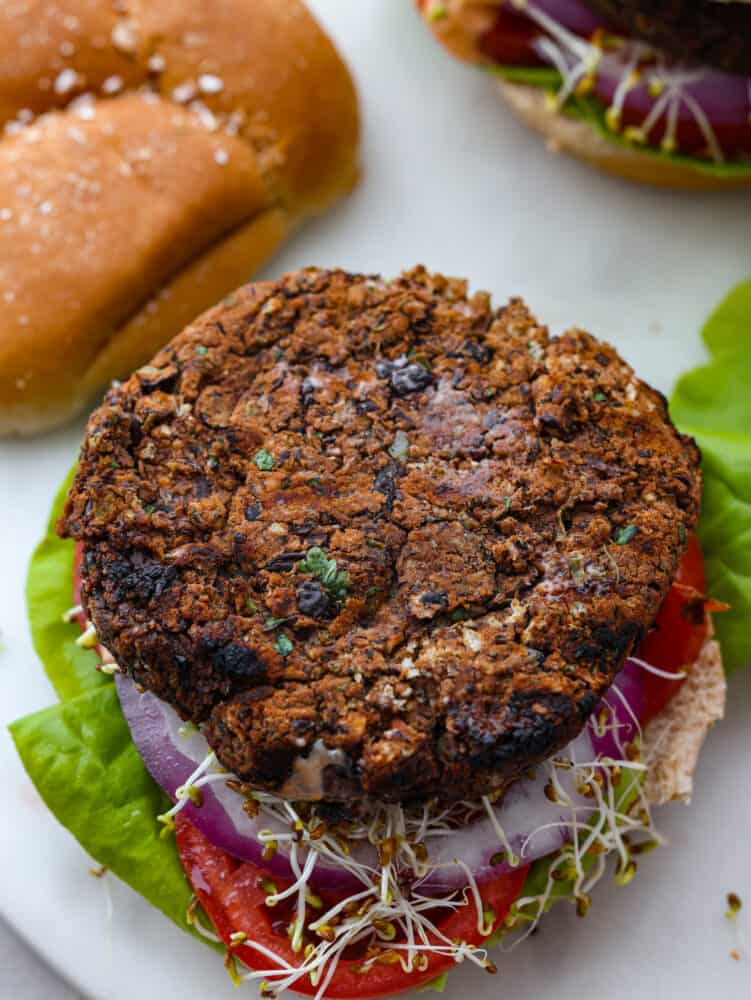 Top view of a black bean burger layered on top of a bun, lettuce, red onion, tomato slice, and sprouts. The top of the bun is removed.