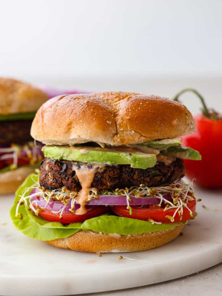 A side view photo of a black bean patty layered in a bun with avocado, tomato, red onion, sprouts, lettuce, and dripping burger sauce.