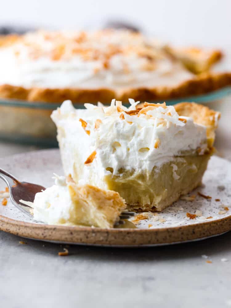 A slice of coconut cream pie on a plate with a bite being cut out with a fork. 