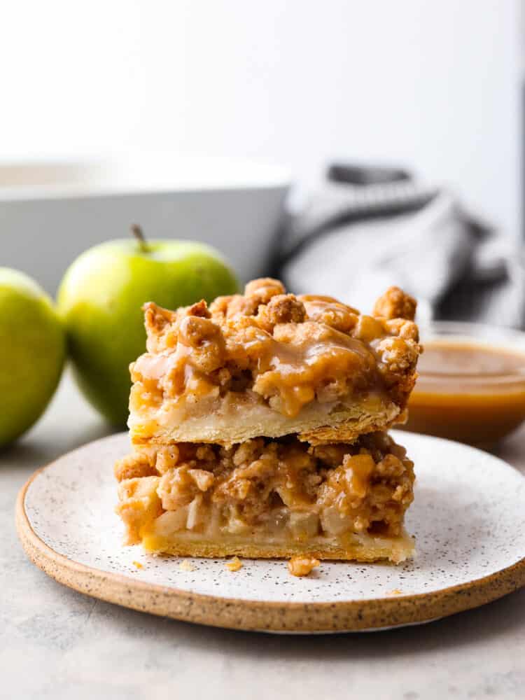 A stack of 2 apple pie bars on a plate with some granny smith apples in the background. 