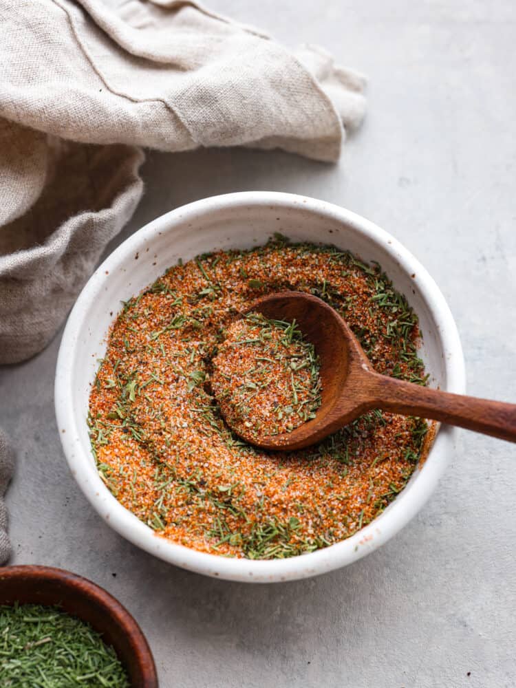 Overhead photo of fish seasoning in a cream bowl with a wood spoon scooping seasoning up. A beige bowl and wood bowl filled with dried parsley are next to the seasoning on a gray countertop.