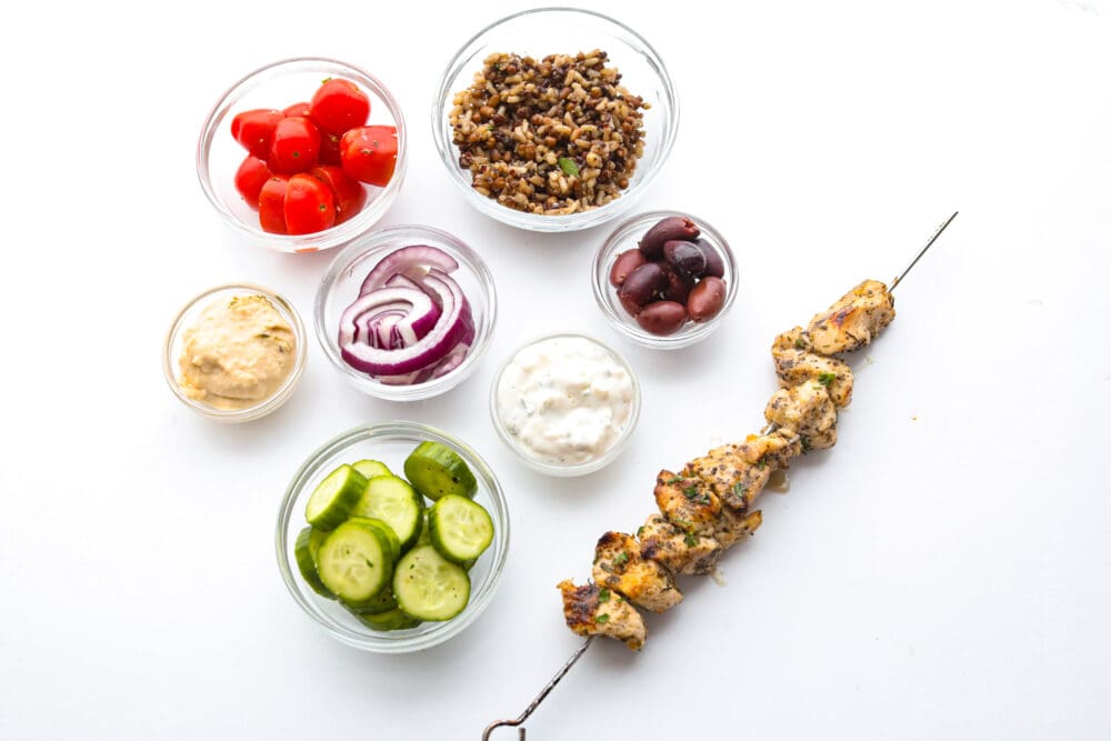 Photo of Greek chicken bowl ingredients in individual bowls.  Also pictured is grilled chicken souvlaki on a skewer.