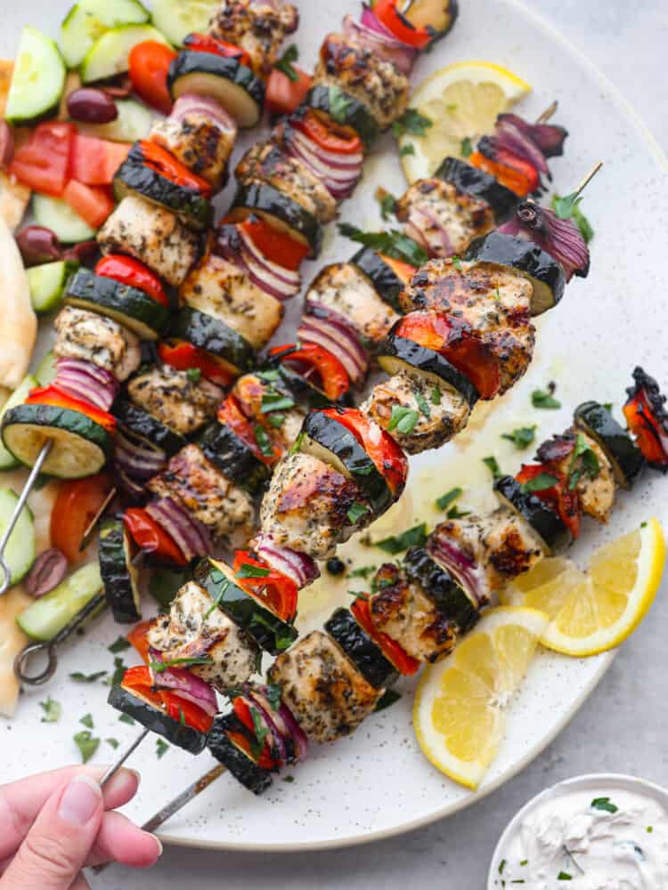 Close up photo of Greek chicken kabobs on a large white round platter. Sliced pita bread, sliced cucumbers, diced tomatoes, and kalamata olives served on the side. Sliced lemon garnished on the side with a small bowl of tzatziki sauce. A hand is holding up one of the kabobs.