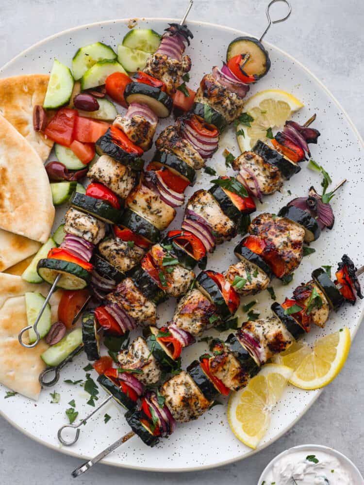Overhead photo of Greek chicken kabobs on a large white round platter.  Sliced pita bread, sliced cucumbers, diced tomatoes, and kalamata olives served on the side.  Sliced lemon garnished on the side with a small bowl of tzatziki sauce.