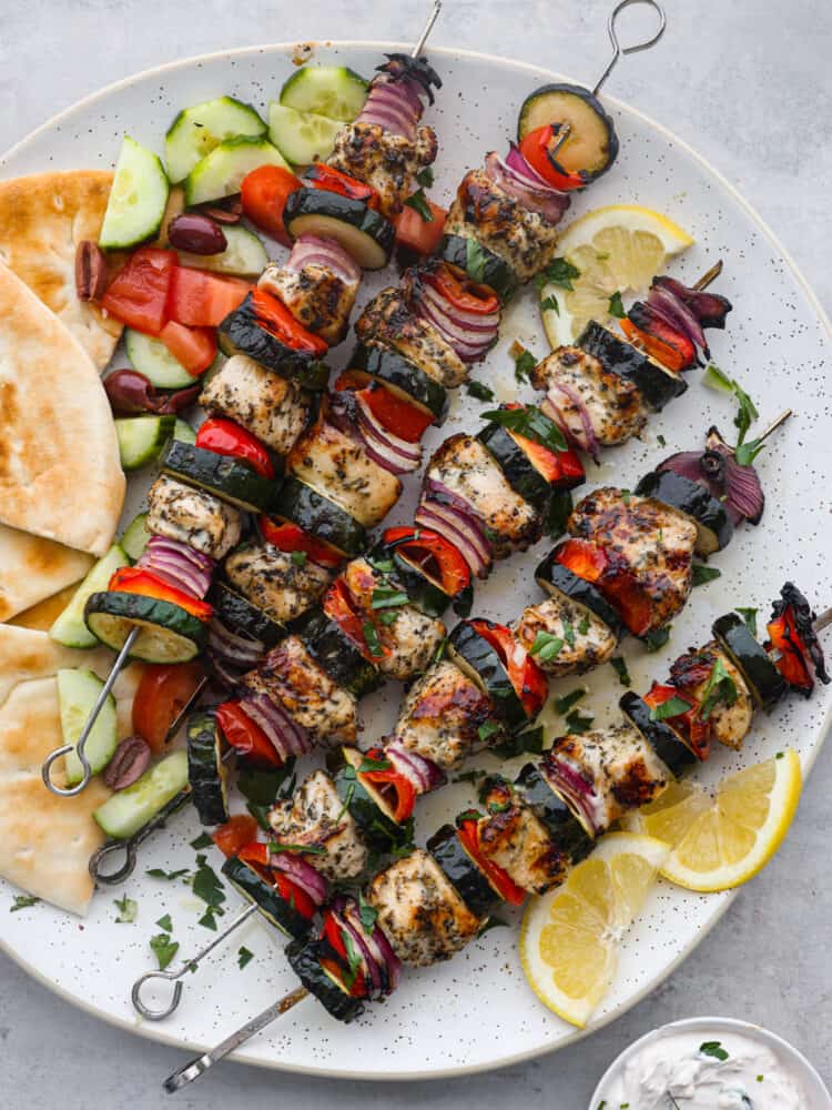 Overhead photo of Greek chicken kabobs on a large white round platter. Sliced pita bread, sliced cucumbers, diced tomatoes, and kalamata olives served on the side. Sliced lemon garnished on the side with a small bowl of tzatziki sauce.