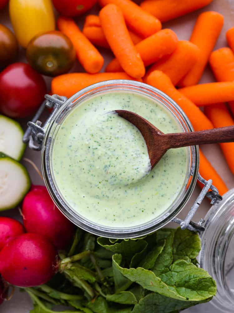 Close up photo of green goddess dressing in a small glass container. A wood spoon is lifting up a spoon full of dressing out of the jar.  Carrots, grape tomatoes, sliced zucchini, and whole radishes scattered around the dressing.