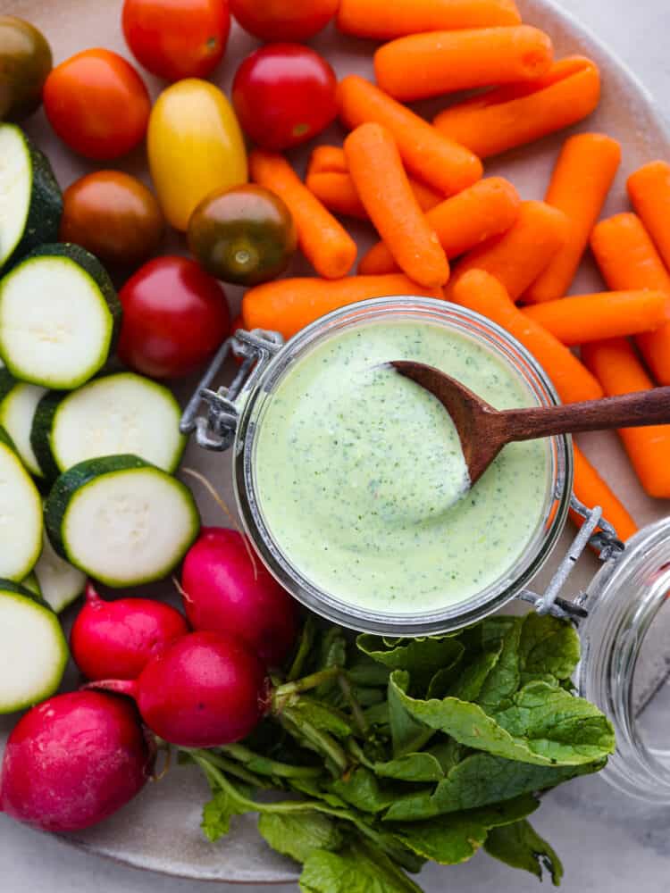 Top view of green goddess dressing in a small glass container with a wood spoon in the jar.  Carrots, grape tomatoes, sliced zucchini, and whole radishes scattered around the dressing.