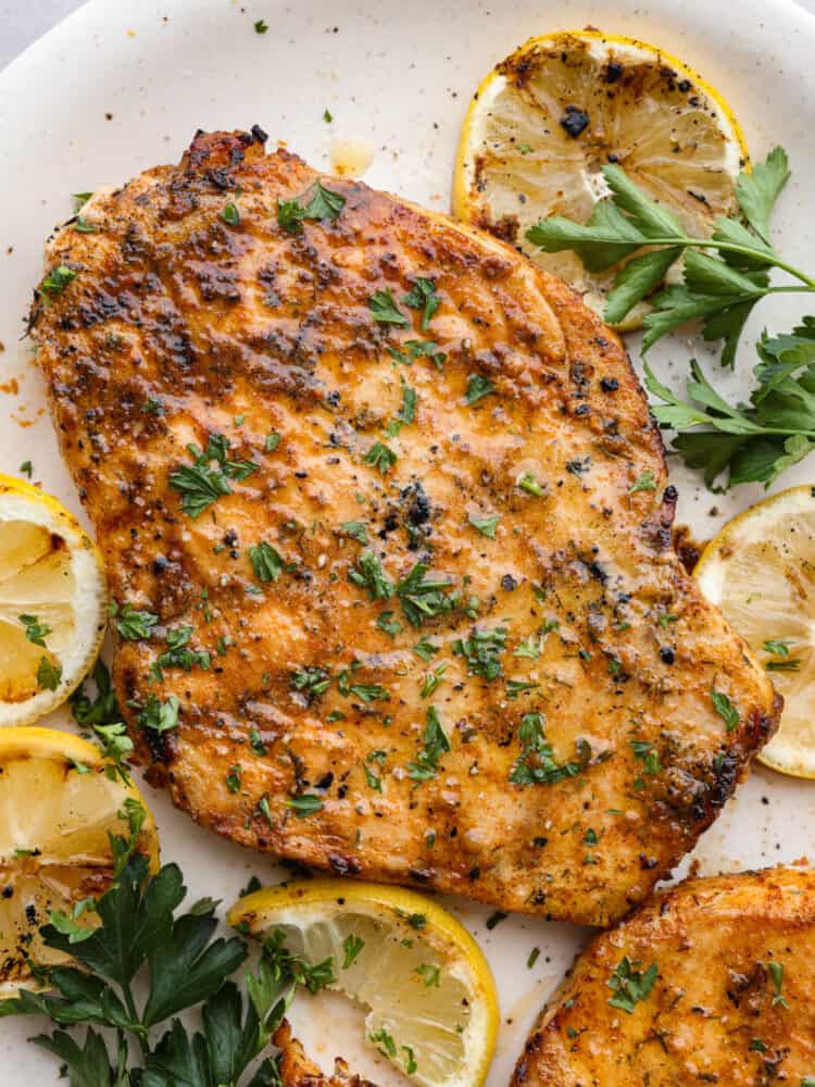 Close up view of grilled swordfish on a white platter. The swordfish is garnished with parsley and grilled lemon slices.