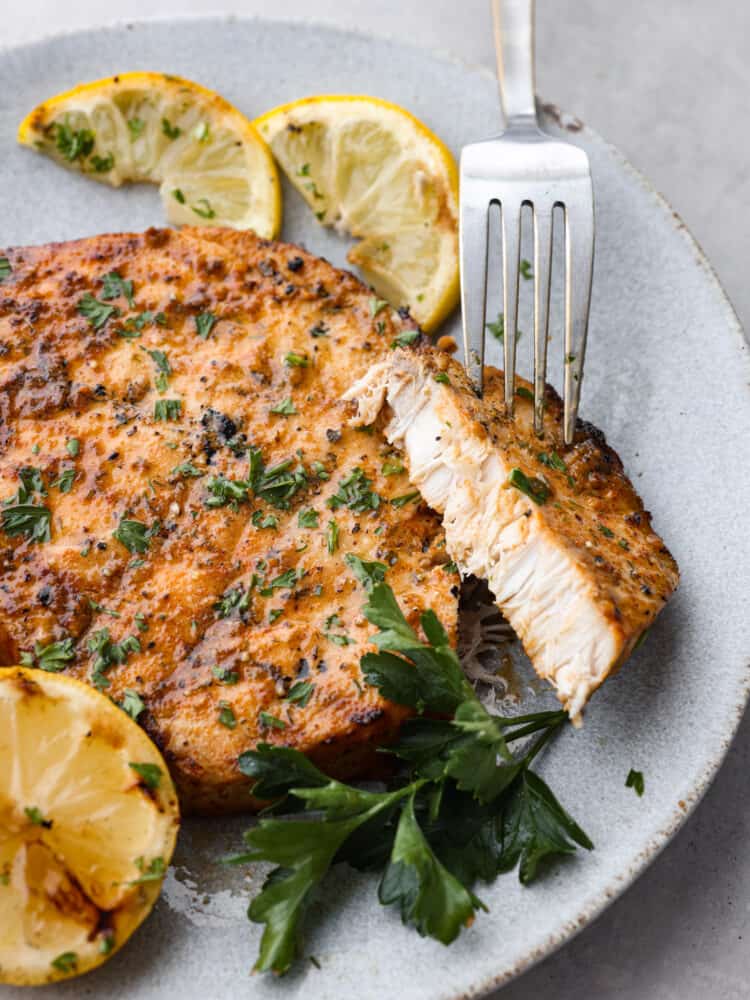 Close up photo of grilled swordfish with a fork lifting up a cut piece.  Garnished with grilled lemon wedges and parsley leaves.