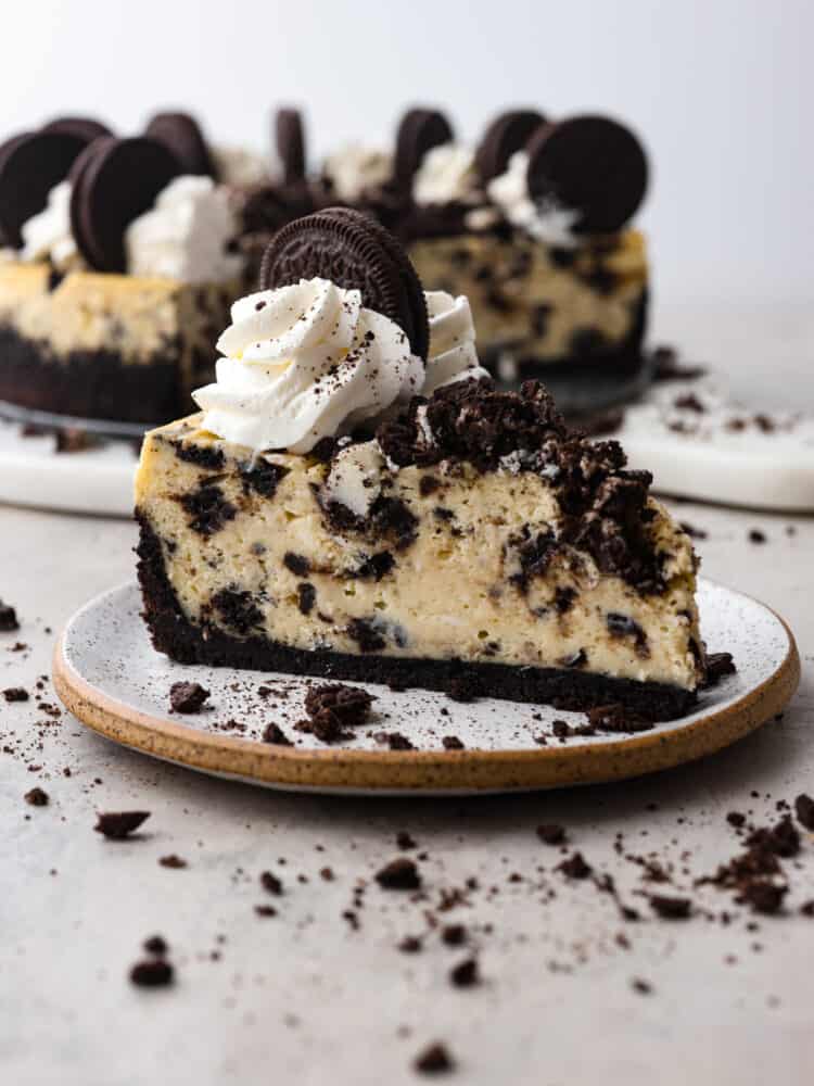 A slice of oreo cheescake on a plate with some oreo crumbs sprinkles around it. 