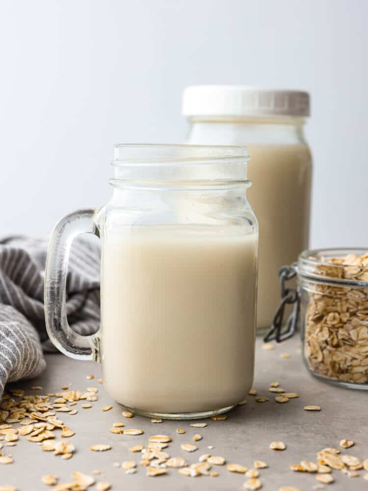 A glass jar with a handle filled with homemade oat milk. There are oats scattered around it.