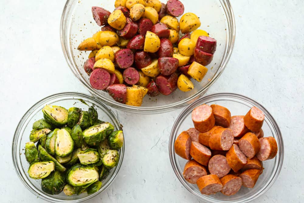 A picture showing 3 glass bowls. One has cut up potatoes, one has sliced Brussel sprouts and the other has sliced sausage. 