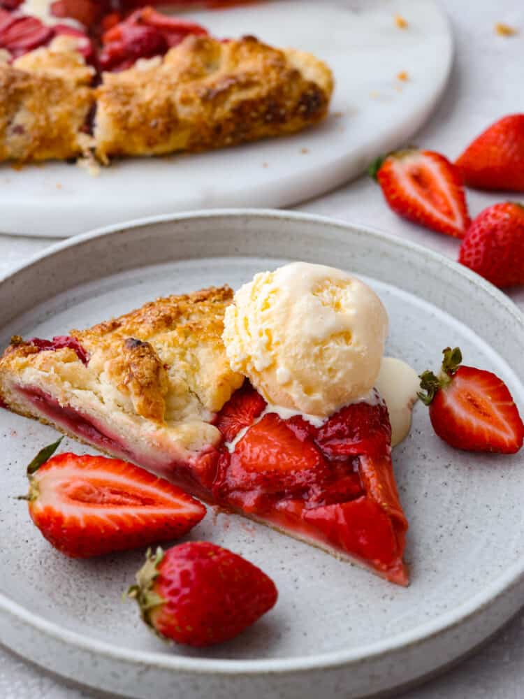 A slice of strawberry galette on a gray plate topped with a scoop of ice cream, and garnished with fresh strawberries on the side.  A view of the remaining galette is pictured in the distance.