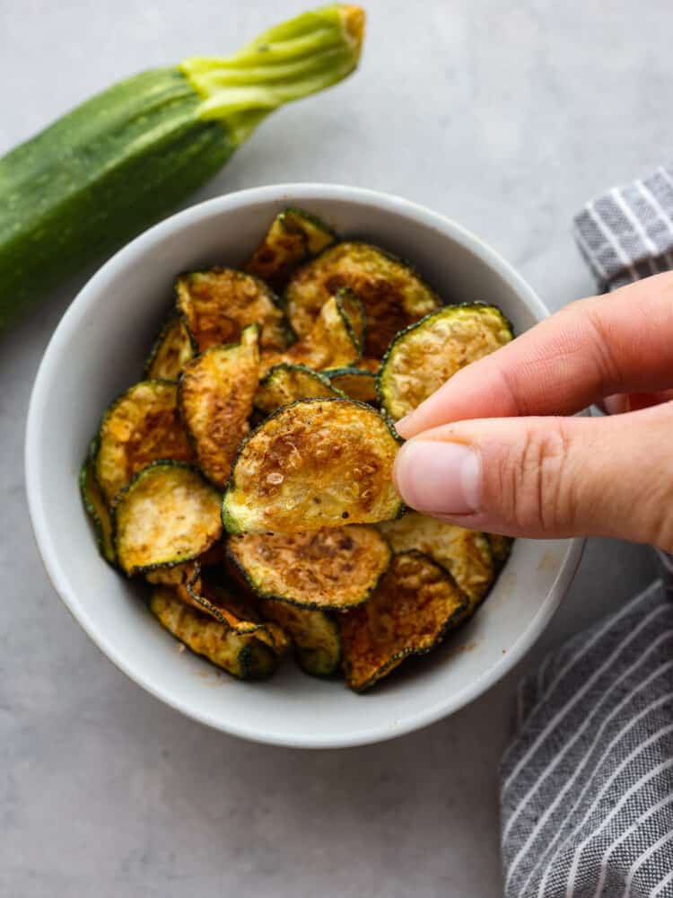 A zucchini chip being picked up out of a small white bowl.