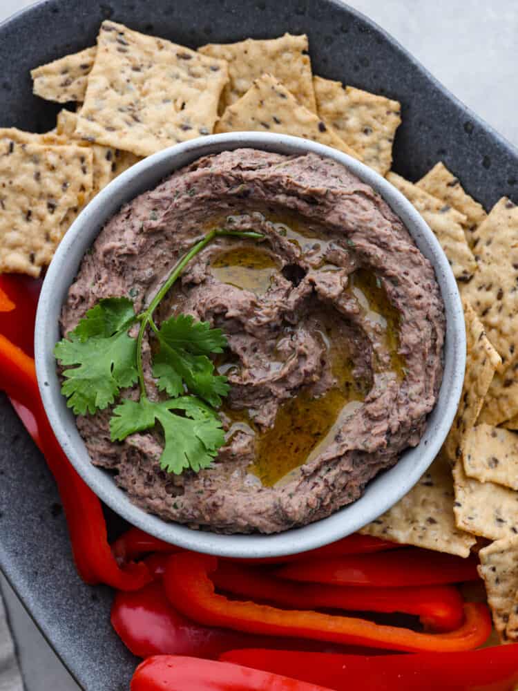 Close up photo of black bean hummus in a gray bowl garnished with olive oil and cilantro.  The hummus bowl is on a gray speckled platter with red peppers and flaxseed crackers surrounding the dip.