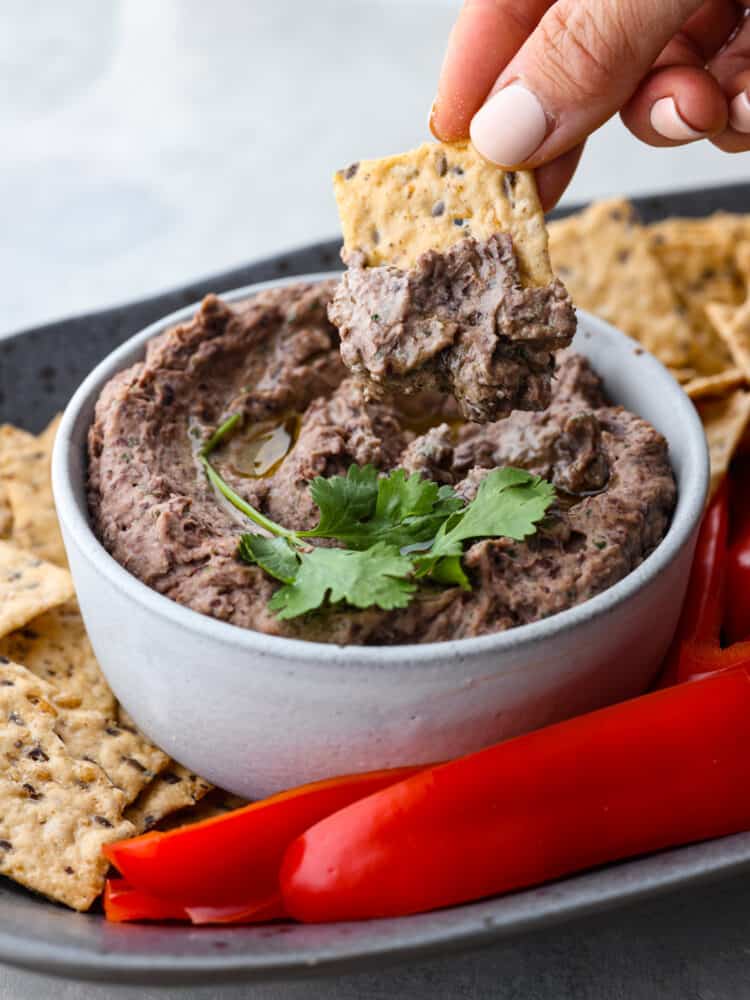 Close up photo of black bean hummus in a gray bowl garnished with olive oil and cilantro.  A cracker is being dipped into the hummus.  The hummus is on a gray speckled platter with red peppers, flaxseed crackers, and celery surrounding the dip.