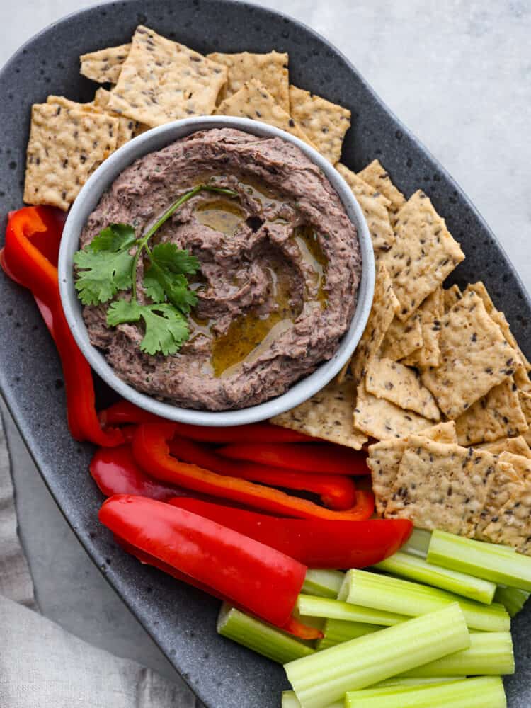 Overhead photo of black bean hummus in a gray bowl garnished with olive oil and cilantro.  The hummus is on a gray speckled platter with red peppers, flaxseed crackers, and celery surrounding the dip.