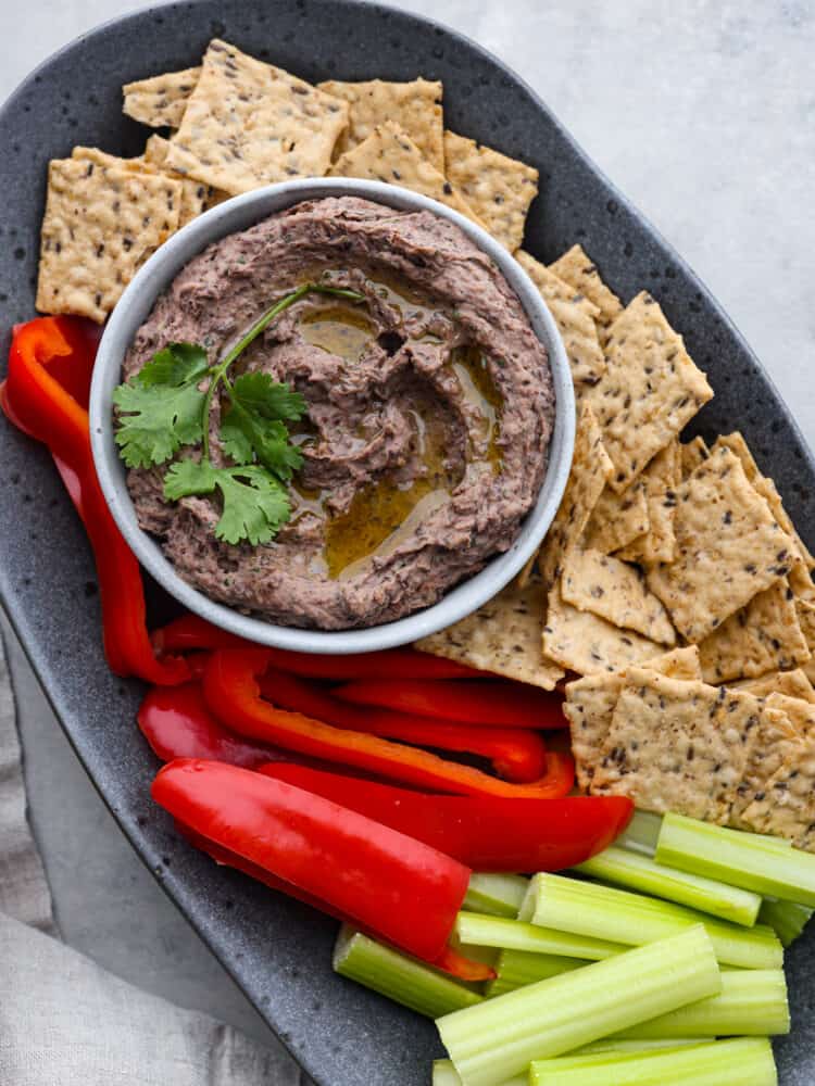 Overhead photo of black bean hummus in a gray bowl garnished with olive oil and cilantro. The hummus is on a gray speckled platter with red peppers, flaxseed crackers, and celery surrounding the dip.