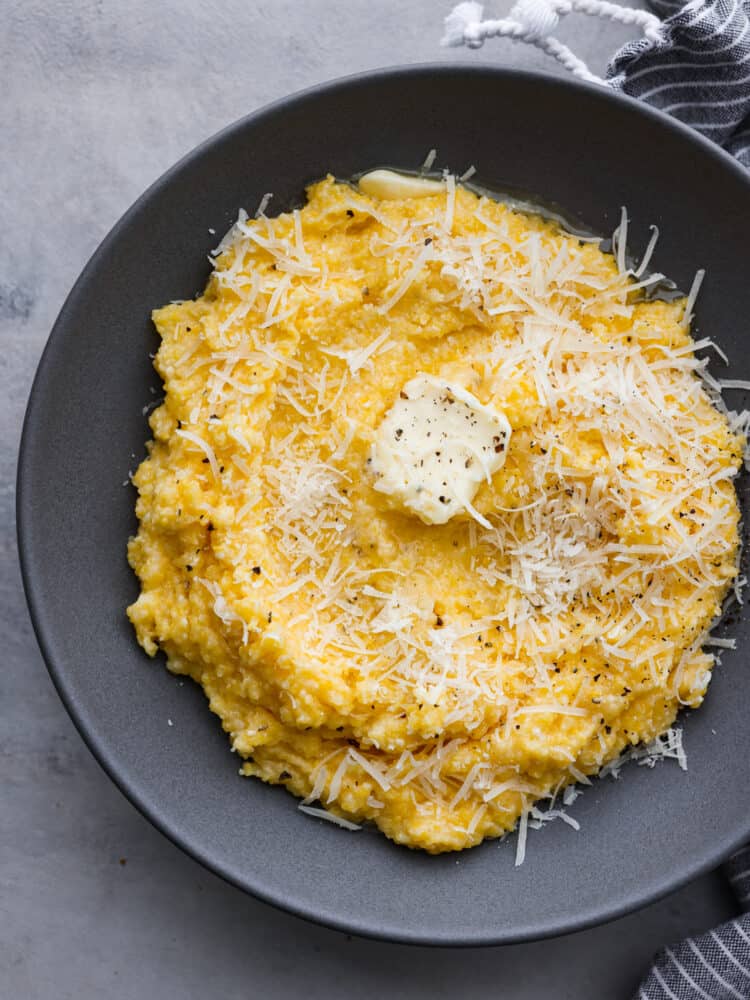 Creamy polenta in a gray bowl with butter, shredded parmesan cheese, and cracked black pepper garnished on top.  A striped gray and white kitchen towel is next to the bowl of polenta.