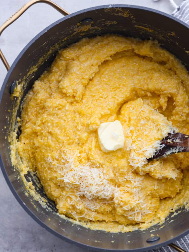 Overhead photo of creamy polenta in a large gray pot.  A pad of butter and shredded parmesan cheese are garnished on top.  A wooden spoon is also sitting in the cooked polenta.