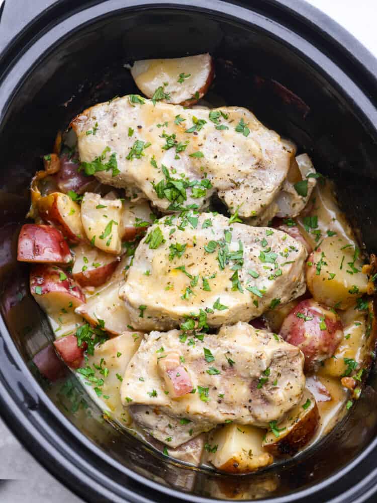 Cooked pork chops and potatoes covered in a creamy, golden sauce garnished with parsley in the base of a slow cooker.