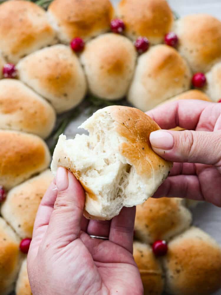 Two hands ripping open a dinner roll with the full dinner roll wreath in the background.