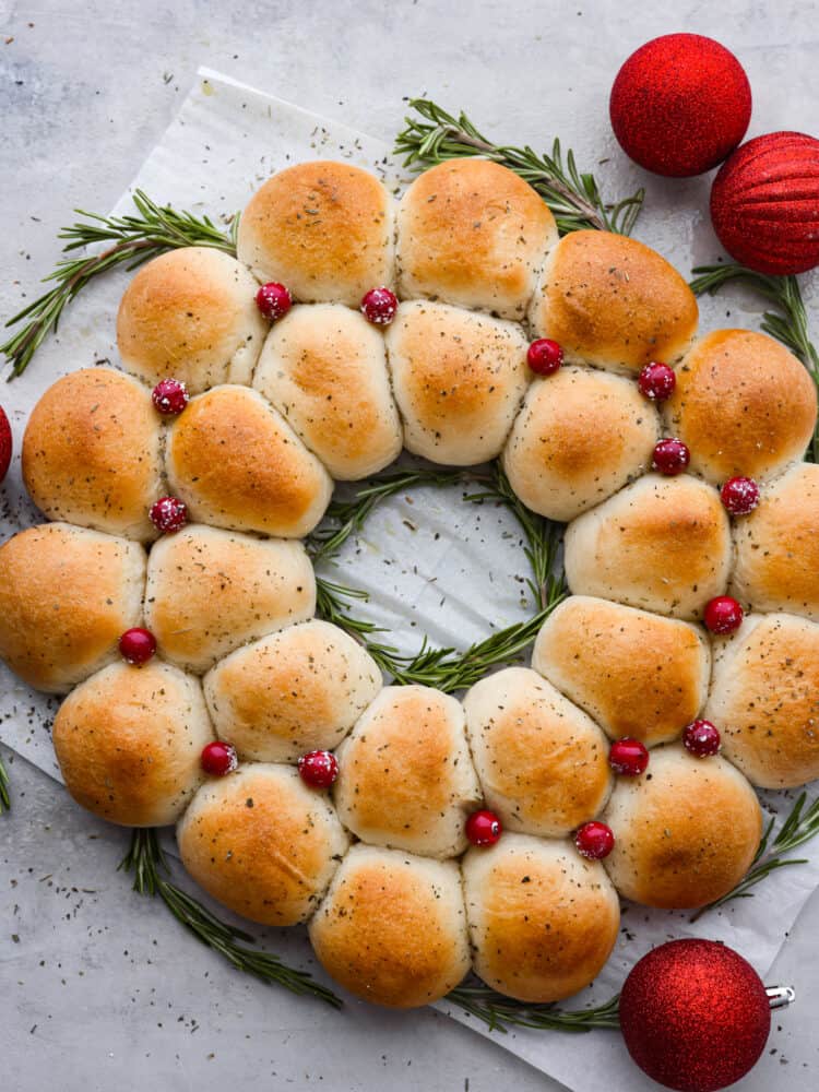 Dinner rolls arranged like a wreath with cranberries on top and rosemary sprigs to the side and red sparkly ornaments.