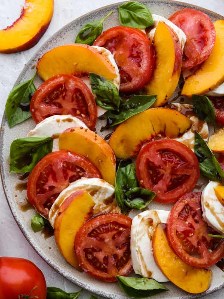 Top-down view of sliced ​​peaches, tomatoes, mozzarella, and basil on a gray plate, topped with balsamic drizzle.