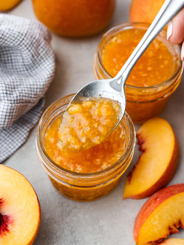 Two small glass jars of peach jam surrounded by a kitchen towel, and sliced peaches.  A spoon is lifting out a spoonful of jam.