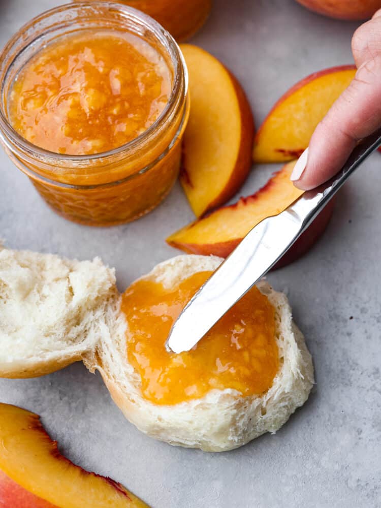 A small glass jar of jam surrounded by sliced peaches an an open faced roll.  A small knife is spreading jam on the roll.