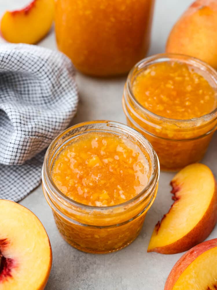 Two small glass jars of peach jam surrounded by a kitchen towel, sliced peaches, and a large glass jar of jam.