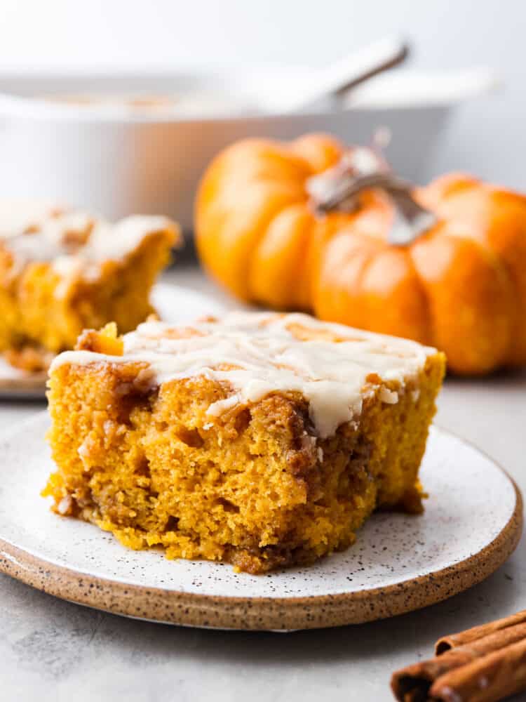 A slice of 0umpkin cinnamon roll cake on a speckled plate with small pumpkins in the background of the picture. 