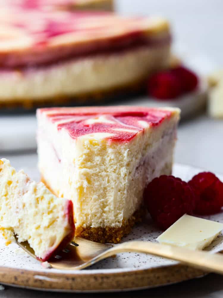 The side view of a slice of white chocolate raspberry cheesecake on a plate with a fork that cut a bite out. 