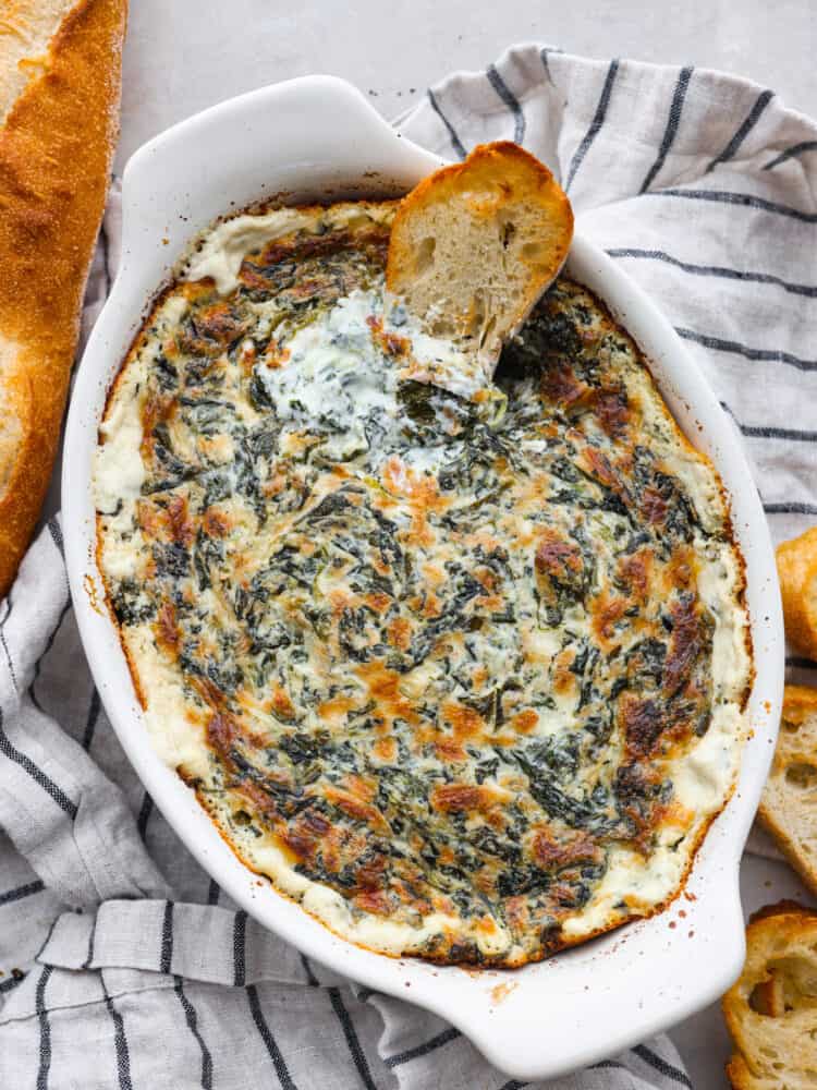 Top view of spinach dip in a oval white baking dish.  A striped dish towel, baguette, and baguette slices are garnished on the side.  A baguette slice is dipped in the spinach dip.