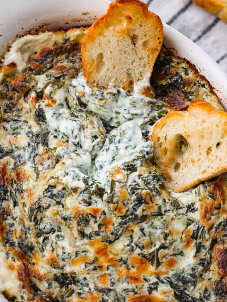 A close up photo of the baked spinach dip and two baguette slices dipping into the dip.