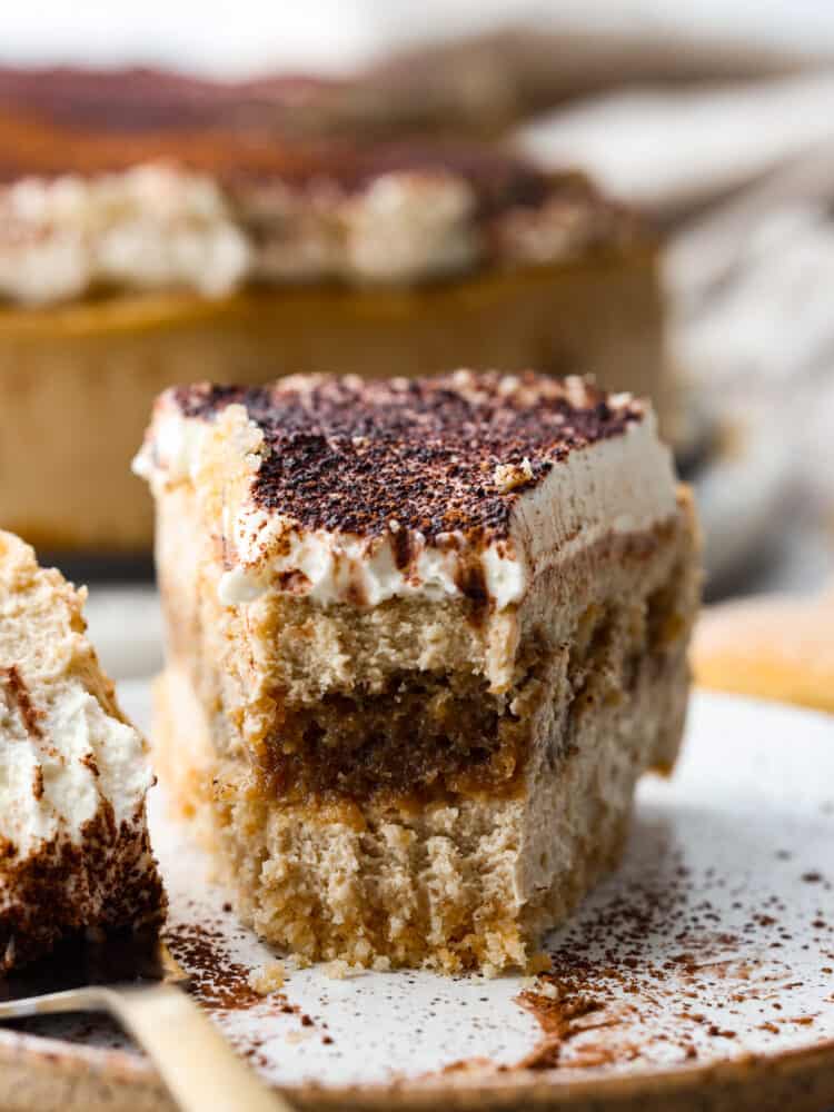 A slice of tiramisu cheesecake on a plate with a bite taken out of it. 
