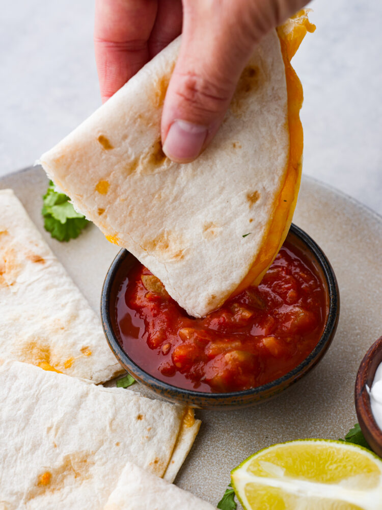 Close up photo of a piece of quesadilla being dipped into a small bowl of salsa.  The quesadilla and salsa are on a large gray plate.  Cilantro and a lime wedge are garnished on the plate.