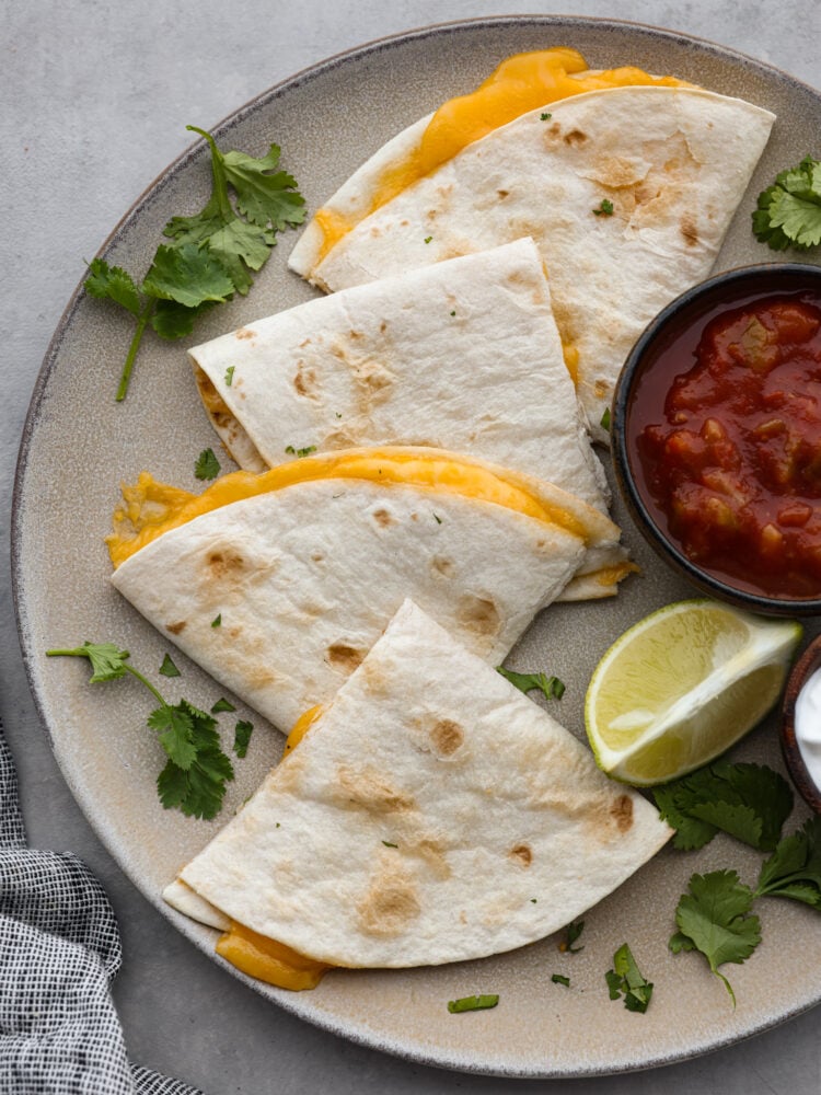 Top view of an air fryer quesadilla cut into triangles on a large gray plate. A small bowl of salsa, cilantro leaves, and a lime wedge are also on the plate next to the quesadilla.