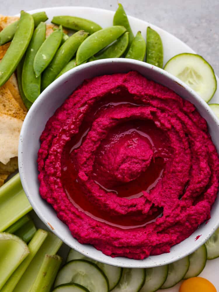 Top close up view of beet hummus in a gray bowl on top of a white marble cutting board.  Vegetables and pita chips are scattered around the hummus.