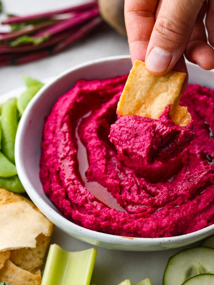 Close up view of beet hummus in a gray bowl. A pita chip is being dipped in the hummus with olive oil garnished on top. Vegetables are served on the side of the dip.
