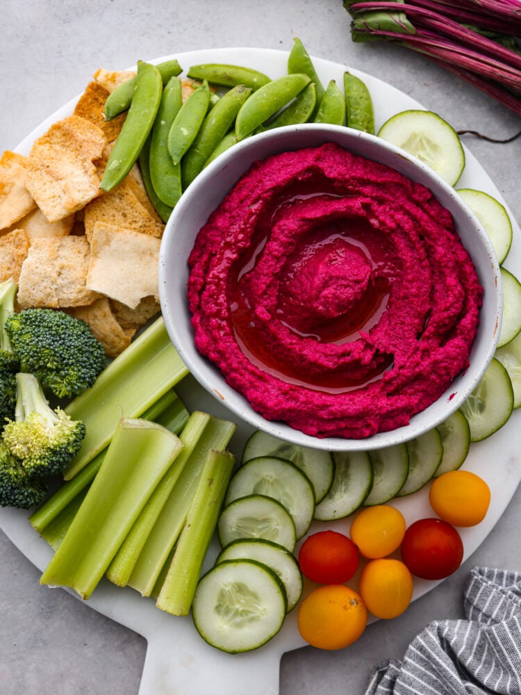Top view of beet hummus in a gray bowl on top of a white marble cutting board.  Vegetables and pita chips are scattered around the hummus.
