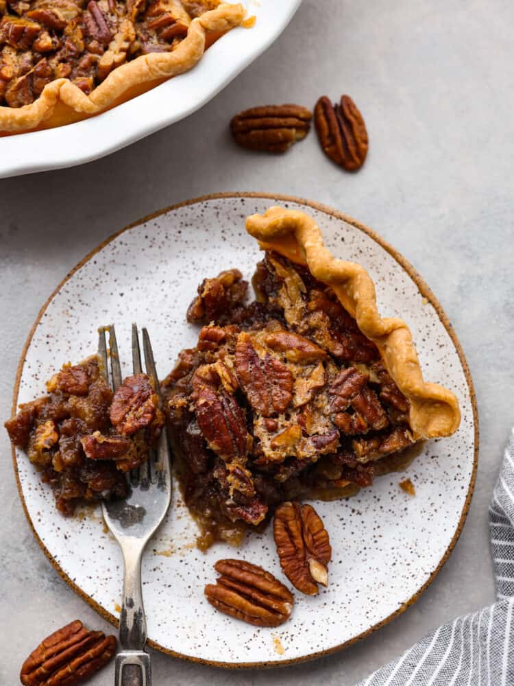 A slice of pecan pie with a bite taken out sitting on a fork on a stone like plate with the rest of the pie to the side.