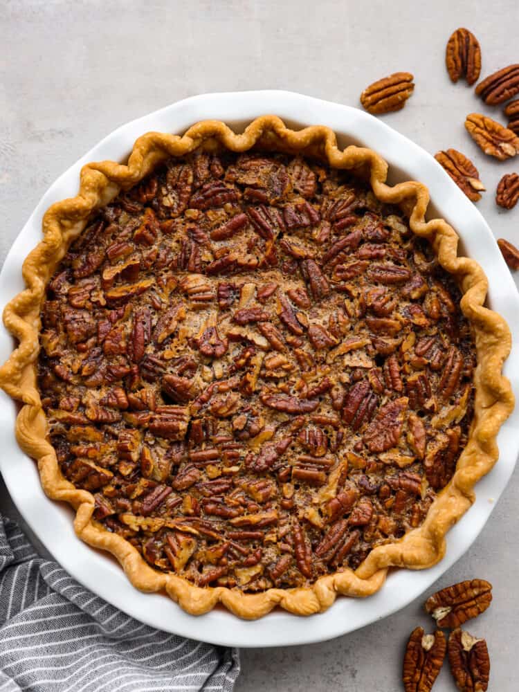 A top view of a whole Bourbon Pecan Pie  with a striped tea towel under neath and pecans sprinkled to the side.