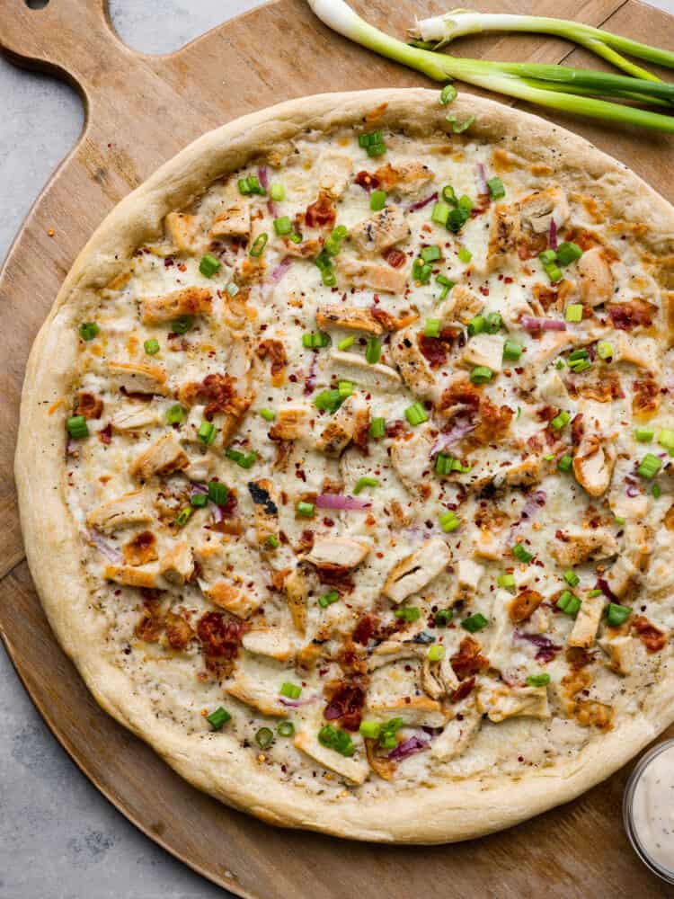 A whole chicken alfredo pizza on a wooden serving board.