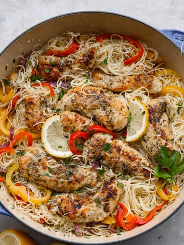 Top-down view of breaded chicken, angel hair pasta, and chopped veggies in a white skillet, garnished with lemon.