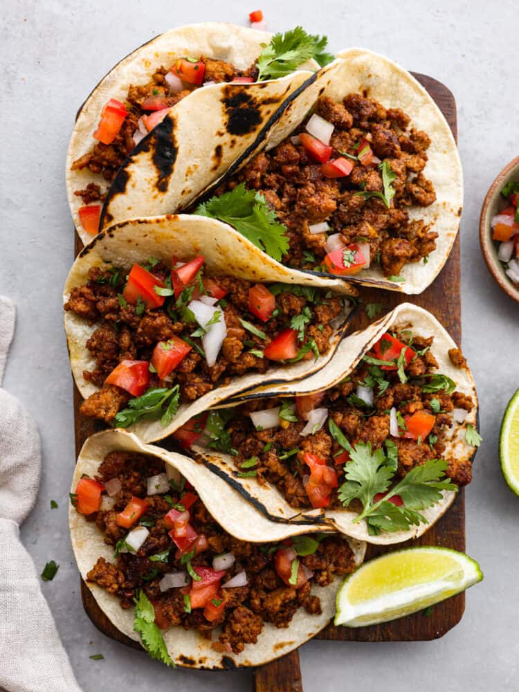 Five Chorizo tacos sitting on a cutting board with tomatoes and onions and cilantro on top with limes to the side.