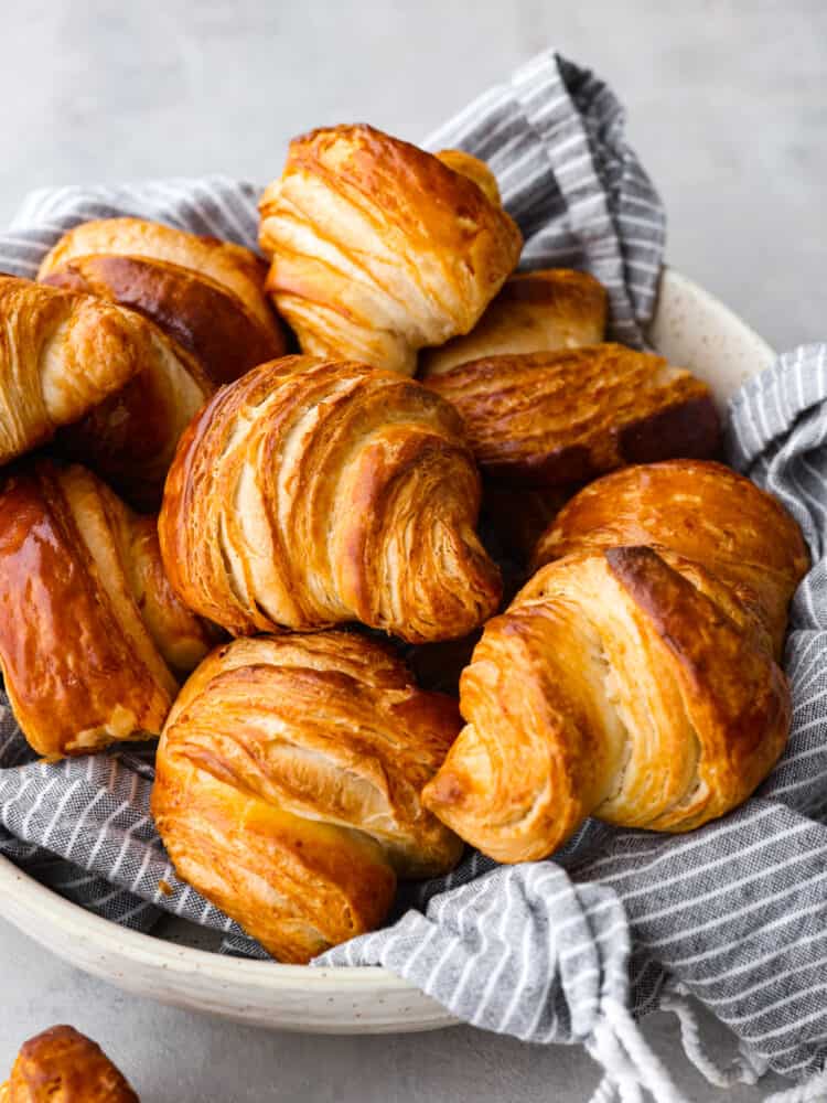 Close up view of croissants stacked on a gray striped kitchen towel and in a large cream colored bowl.