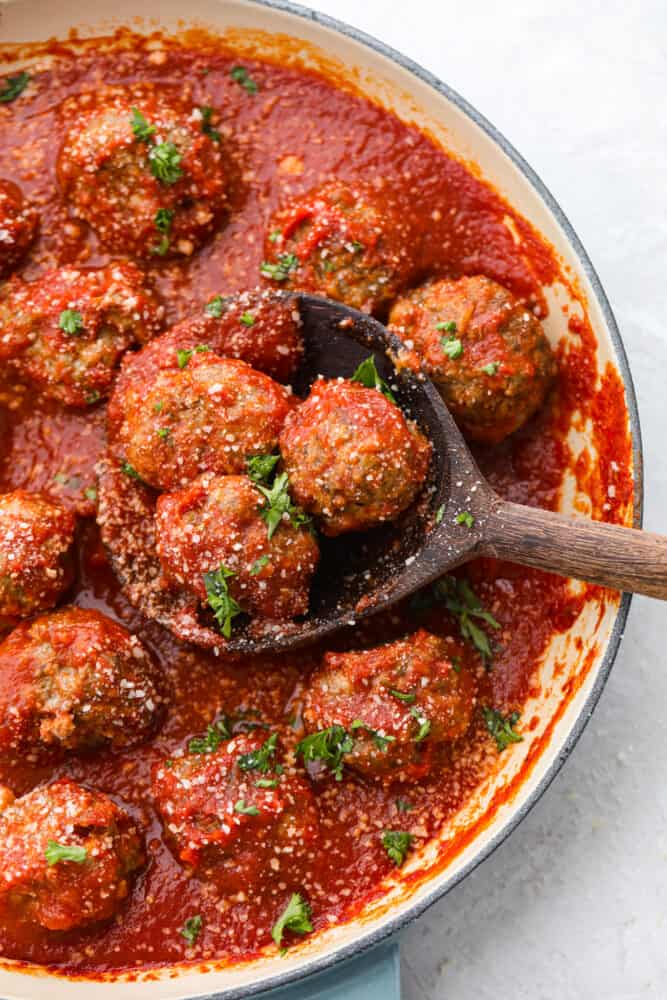 3 meatballs covered in marinara sauce being scooped out of a skillet with a wooden spoon.