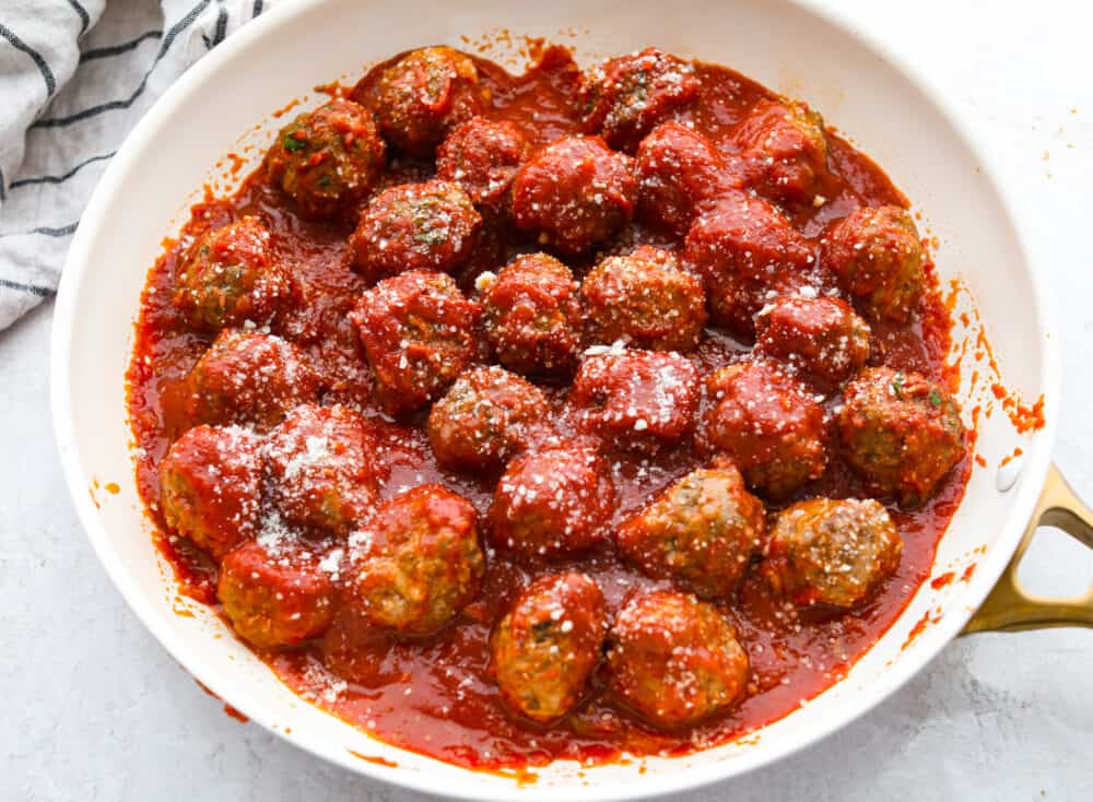 Cooked meatballs in marinara sauce, topped with grated parmesan in a white skillet.